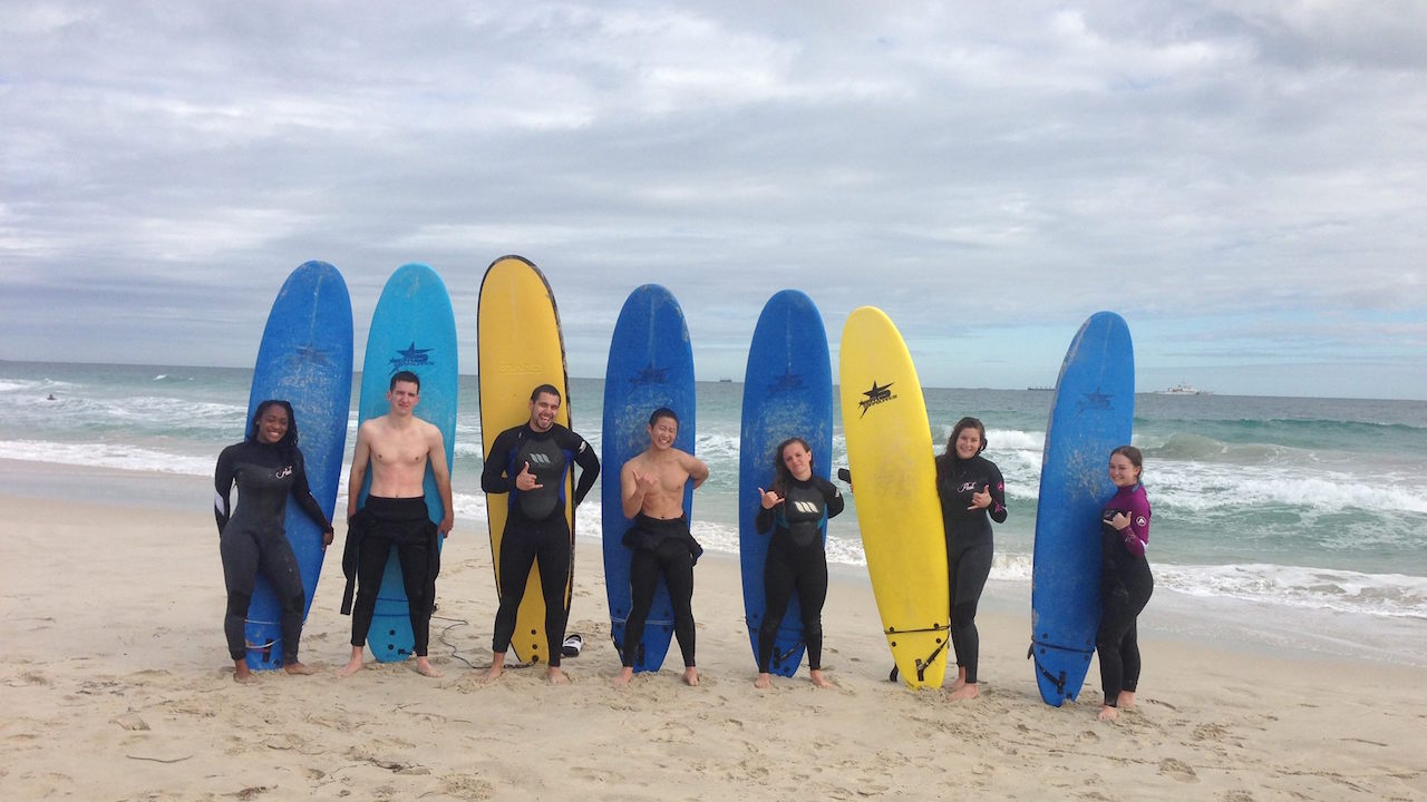 Seven students pose with their surf boards on the beach under a cloudy sky