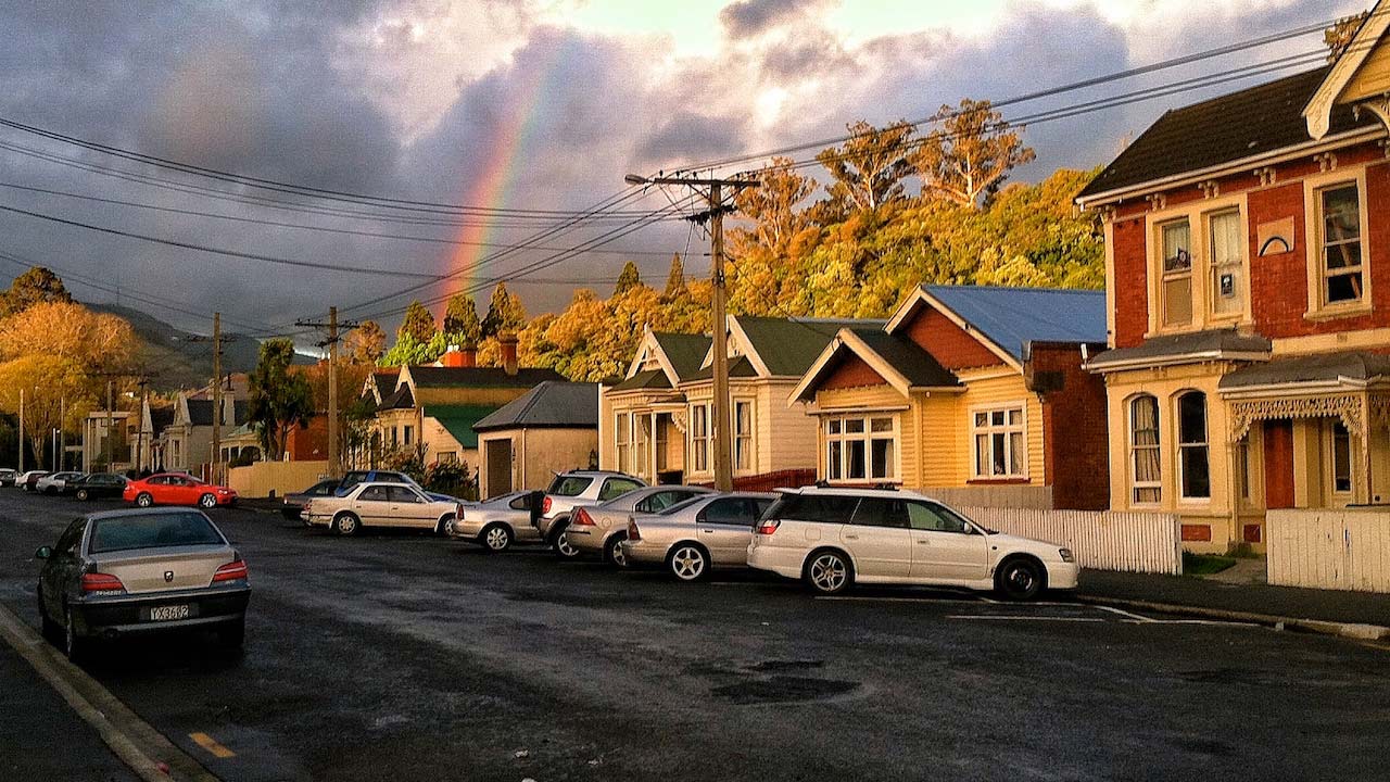 A rainbow peeks out of the cloudy sky behind a row of homes and parked cars in Dunedin, New Zealand