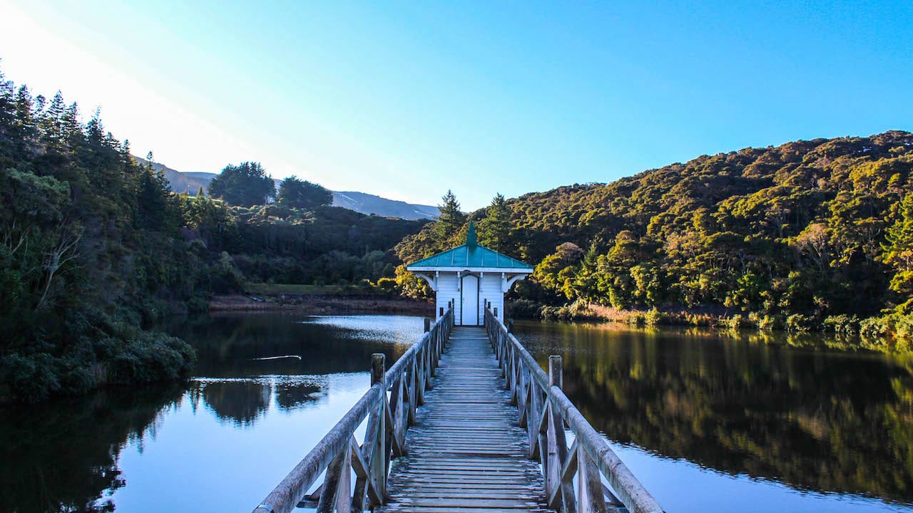 A wooden bridge leading to a house on a crystal clear lake surrounded by mountains near Dunedin, New Zealand
