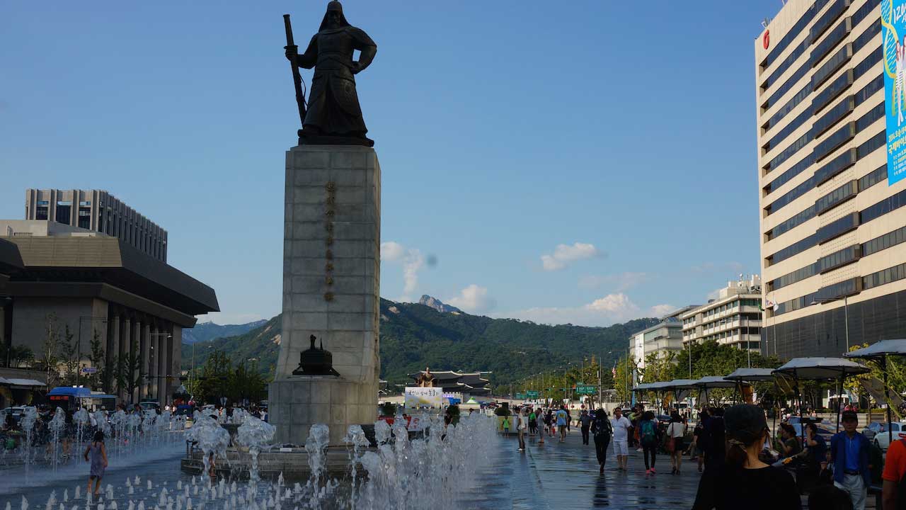 People walk around fountain and a statute surrounded by buildings in Seoul