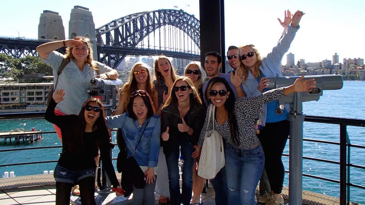 A group of students pose enthusiastically in front of the Sydney Bridge