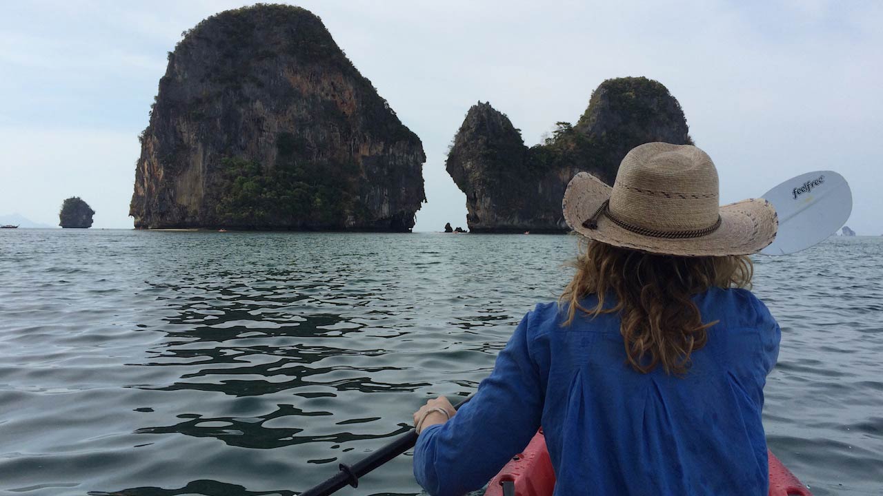 The back of a girl paddling a kayak in the ocean towards limestone cliffs in Railay, Thailand