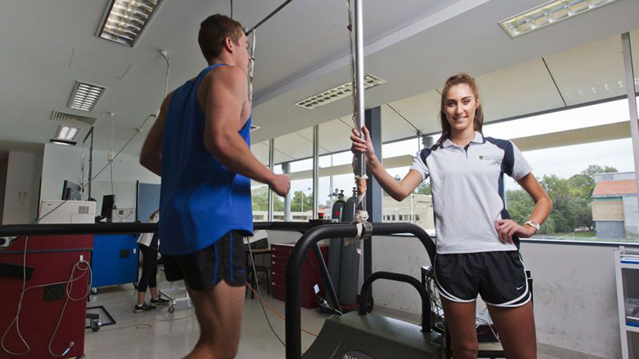 A woman stands smiling next to a man exercising in a classroom at University of Western Australia in Perth